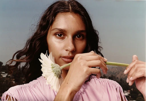Close-up portrait of a young woman holding a daisy, with an urban hillscape and bright sky providing a background in soft light.