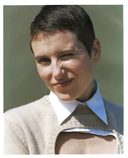 A woman with short hair, wearing a cream-colored cropped sweater and white collared shirt, winks at the camera in an outdoor setting.