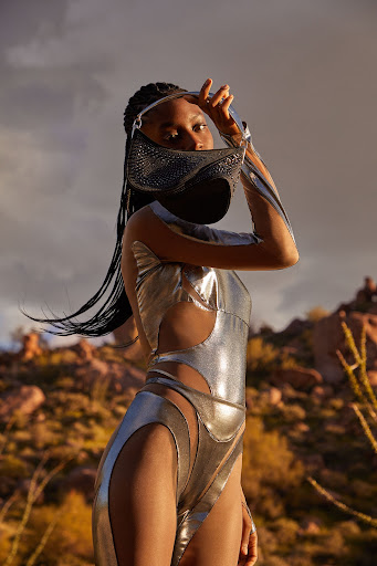 A Black female model in a futuristic silver bodysuit poses outdoors against a desert landscape, holding a jeweled mask to her face.