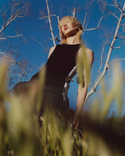 Low angle portrait of a person with short blonde hair wearing a black top, standing outdoors among tall grass and bare trees, against a blue sky.