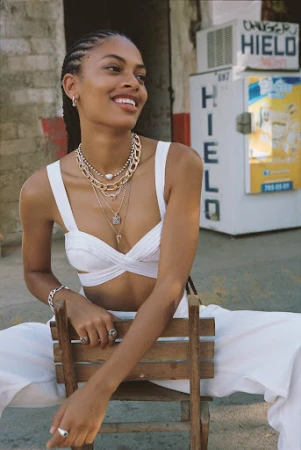 Portrait of a smiling dark-skinned woman with braids in a white top and pants, seated in a chair against an urban background.