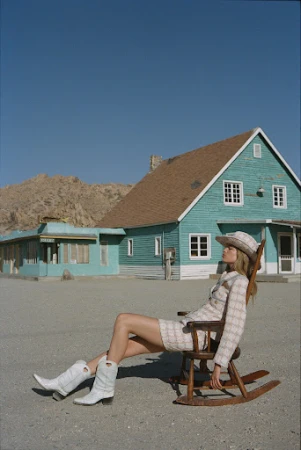 A blonde woman in a cream-colored suit and cowboy boots relaxes in a wooden rocking chair in front of a turquoise desert house against a blue sky.