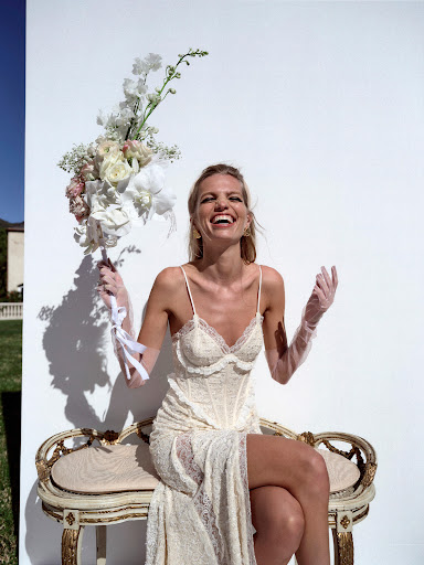 Smiling woman in lace dress sits holding a bouquet of white flowers outdoors against a white wall with greenery in the background.