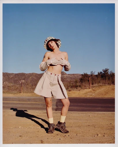 Woman in bucket hat and shorts standing on desert road with mountains and blue sky in the background, casting a long shadow.