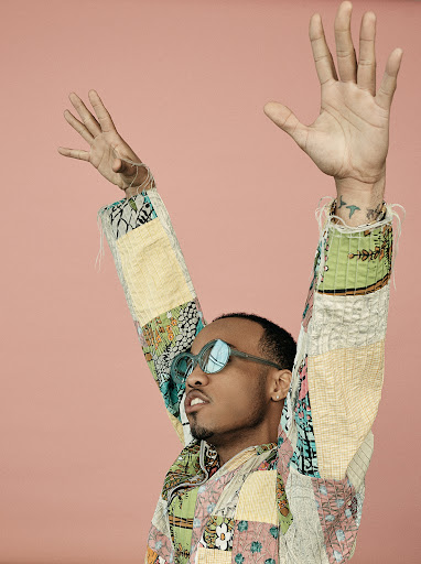 Person with arms raised wearing patchwork jacket and sunglasses against a pink background in a medium studio shot.