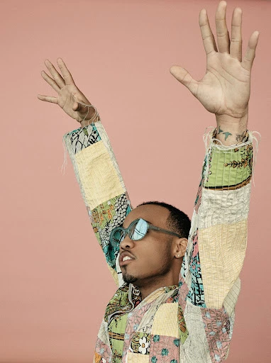 Person with arms raised wearing patchwork jacket and sunglasses against a pink background in a medium studio shot.