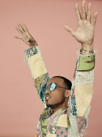 Person with arms raised wearing patchwork jacket and sunglasses against a pink background in a medium studio shot.