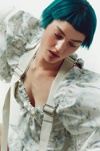 Close-up of a person with short turquoise hair, wearing a floral-print top with suspenders, posing in front of a white background.