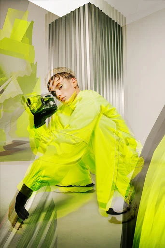 Young adult in neon windbreaker jacket holding vintage camera in creative indoor studio fashion shoot with unique styling.