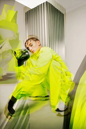 Young adult in neon windbreaker jacket holding vintage camera in creative indoor studio fashion shoot with unique styling.