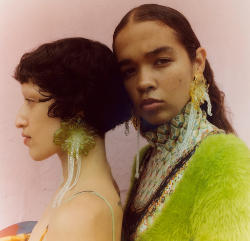 Two young women pose in a portrait wearing artistic green and yellow earrings. One wears a lime-green fur coat and a printed turtleneck.