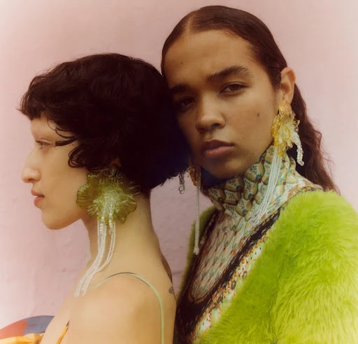 Two young women pose in a portrait wearing artistic green and yellow earrings. One wears a lime-green fur coat and a printed turtleneck.
