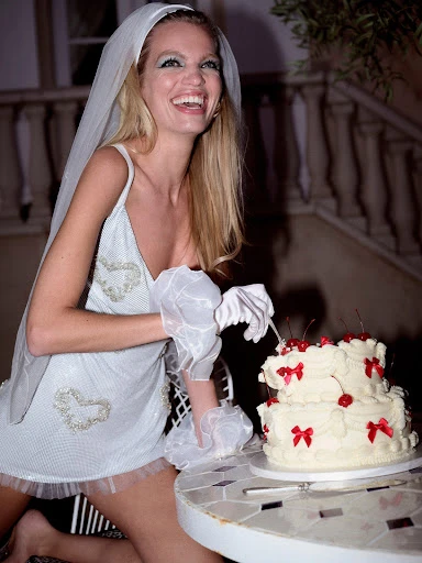 Smiling blond woman in white dress cuts two-tier cake decorated with red cherries and bows; outdoor setting, celebratory mood.