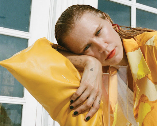 Blonde woman with wet hair poses in patterned shirt and leans on yellow object next to window in indoor setting with soft, diffused light.