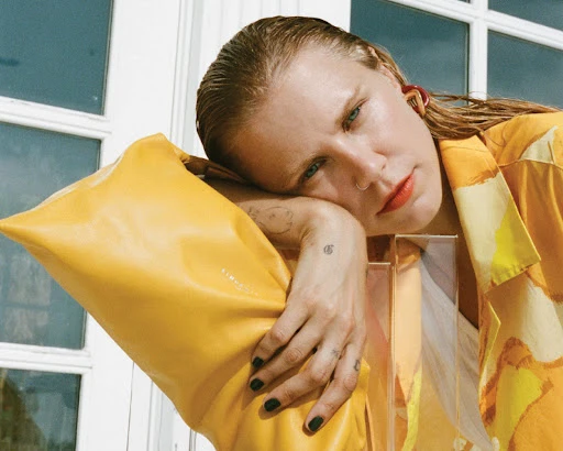 Blonde woman with wet hair poses in patterned shirt and leans on yellow object next to window in indoor setting with soft, diffused light.