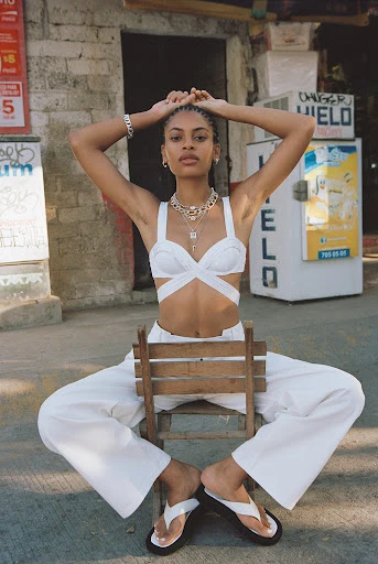 Young woman poses on a wooden chair outdoors wearing a white top, pants, and sandals with a concrete block wall behind her.