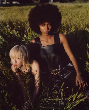 Two female models posing in a tall grassy field with natural sunlight, featuring make-up by Hadia Kabir.