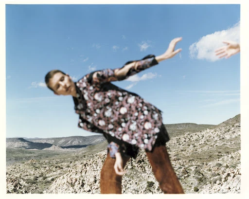 A blurred image captures a person in a floral shirt standing on a rocky outcrop, with another outstretched arm reaching toward them against a sky background.