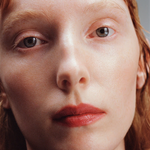 Close-up portrait of a woman with red hair, fair skin, light-colored eyelashes, and subtle red lipstick against a neutral grey background.