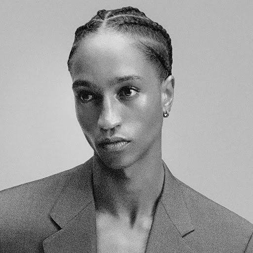 Close-up portrait of an African-American man with cornrow braids wearing a gray suit jacket against a gray backdrop.