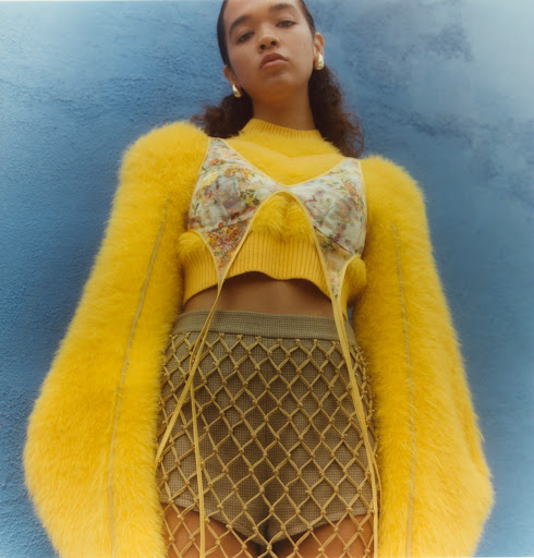 Young adult female poses against blue wall in yellow sweater, floral top, and net skirt. Studio lighting and fashion photography.