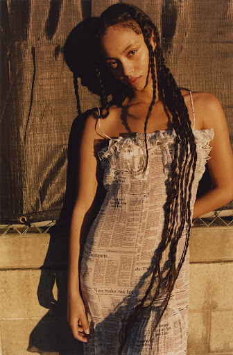 Young woman poses in newspaper-print dress in soft light against textured wall, braids trailing down, editorial fashion shot.