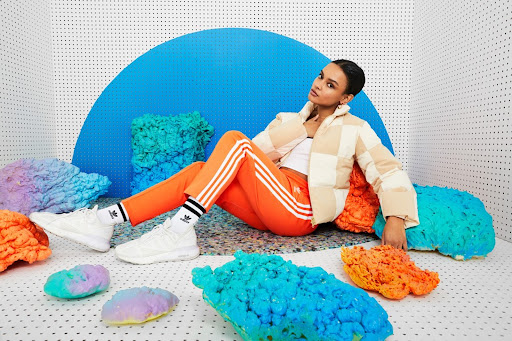 Woman in orange track suit and cream puffer jacket poses in a photo studio with abstract sculptures, promoting fashion and lifestyle.