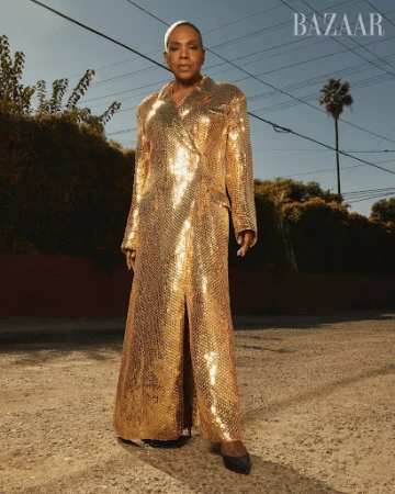 A single adult woman stands outdoors on a road in a gold sequined coat, complemented by black heels, in an eye-level medium shot.