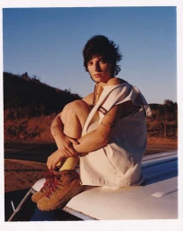 Woman with short dark hair wearing a white sleeveless top and brown hiking boots seated atop a car roof at sunset.