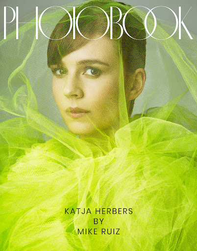 Close-up of a female model with short brown hair wearing a bright yellow tulle top against a gray backdrop for 'Photobook' magazine.