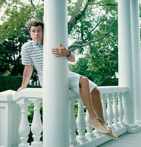 Man leans against a white column outdoors wearing a striped shirt and shorts with tan shoes, green trees visible in the background.