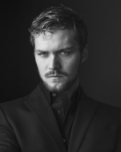 Black and white portrait of an adult male with wavy hair, a mustache, and a beard wearing a collared shirt and blazer against a dark backdrop.