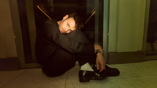 Young man in black suit and white socks sits curled on a green tiled floor indoors with hands clasped in front of a glass door.