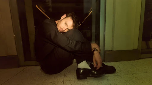 Young man in black suit and white socks sits curled on a green tiled floor indoors with hands clasped in front of a glass door.