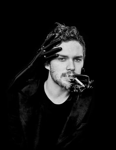 Black and white portrait of a man smoking with hands in black latex gloves framing his face against a dark background.