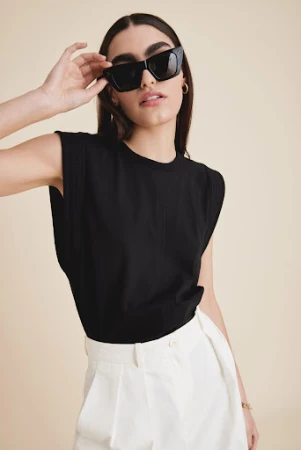 Woman in black sleeveless top and white trousers wearing oversized sunglasses against a neutral background