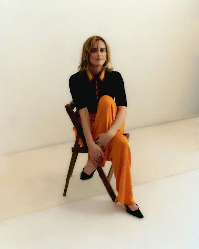 Adult female sitting on a wooden folding chair in a photo studio wearing orange wide-leg pants and a black button-down shirt with yellow trim.