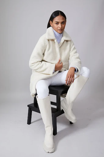 Fashionable woman poses on a stool in a studio wearing a white coat, white pants, and knee-high boots against a neutral background.