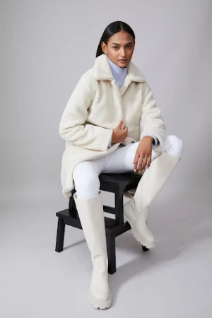 Fashionable woman poses on a stool in a studio wearing a white coat, white pants, and knee-high boots against a neutral background.
