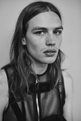 Black and white portrait of a young adult man with long hair, wearing a mesh top with 'DIOR' logo, against a neutral background.