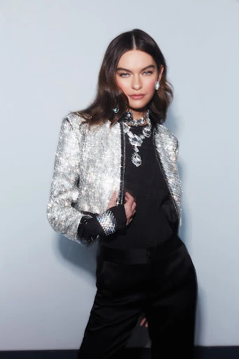 Brunette woman in a sparkling silver jacket and black satin pants, posing against a neutral background, wearing statement jewelry.