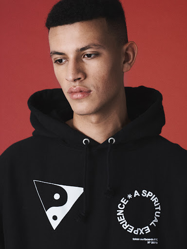 Portrait of a young adult male with short dark hair wearing a black hoodie with a Yin and Yang symbol and printed text against a red backdrop.
