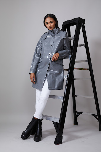 Female model in grey glossy trench coat with white jeans and black boots posing near a black ladder in a studio setting.
