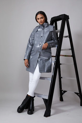 Female model in grey glossy trench coat with white jeans and black boots posing near a black ladder in a studio setting.