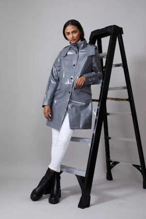 Female model in grey glossy trench coat with white jeans and black boots posing near a black ladder in a studio setting.