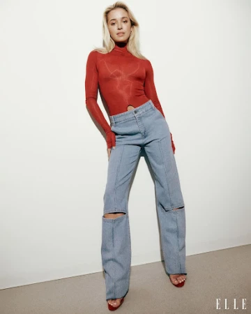 Woman with blonde hair wearing a red turtleneck top and light wash denim jeans with knee cutouts against a white wall.