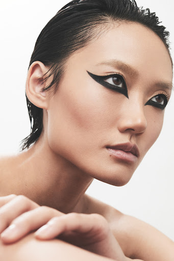 Close-up portrait of a woman with dramatic black winged eyeliner, slicked-back dark hair, neutral makeup, and hands folded near her face.