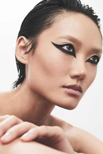 Close-up portrait of a woman with dramatic black winged eyeliner, slicked-back dark hair, neutral makeup, and hands folded near her face.