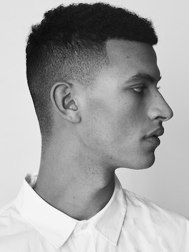 Profile portrait of a young man with short dark hair wearing a white collared shirt against a plain light background