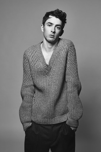 Black and white portrait of a young adult man wearing a wool V-neck sweater and dark pants in front of a neutral grey background.